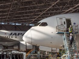 Japan Airlines jet under a metal hangar roof with maintenance workers on a scaffold beside the open door of the forward fuselage