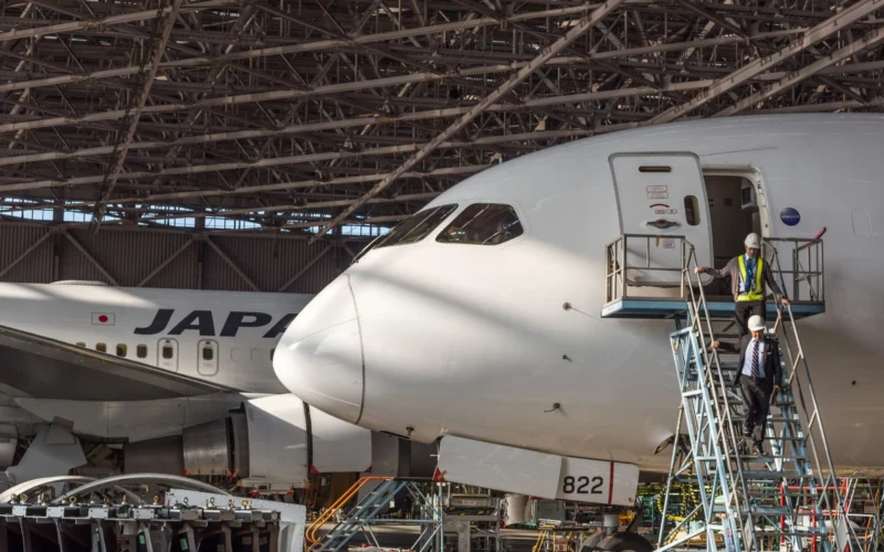 Japan Airlines jet under a metal hangar roof with maintenance workers on a scaffold beside the open door of the forward fuselage