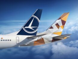 Two commercial aircraft tails in flight one with a blue Lufthansa logo the other with a geometric orange brown pattern against a blue sky