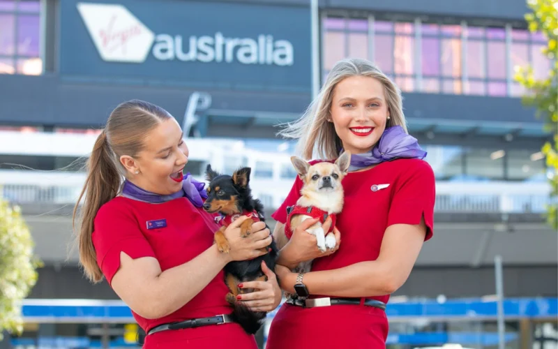 Two women in red uniforms holding small dogs outdoors in front of a building with a visible Australia sign in the background