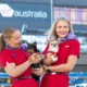 Two women in red uniforms holding small dogs outdoors in front of a building with a visible Australia sign in the background