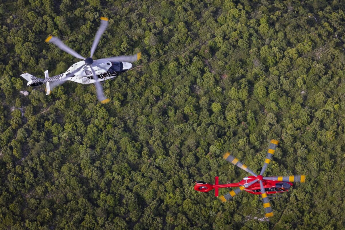 Aerial view of two helicopters a white black marked helicopter in the left center and a red helicopter with yellow rotor tips nearby over a dense forest