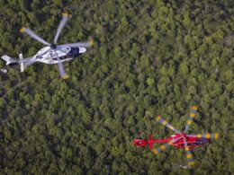 Aerial view of two helicopters a white black marked helicopter in the left center and a red helicopter with yellow rotor tips nearby over a dense forest