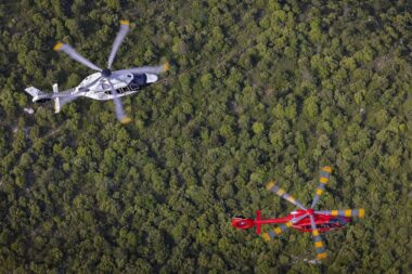 Aerial view of two helicopters a white black marked helicopter in the left center and a red helicopter with yellow rotor tips nearby over a dense forest