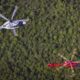 Aerial view of two helicopters a white black marked helicopter in the left center and a red helicopter with yellow rotor tips nearby over a dense forest