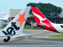 Two airplane tails at an airport one with an orange star design and the other red with a white kangaroo logo parked near a hangar