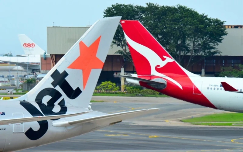 Two airplane tails at an airport one with an orange star design and the other red with a white kangaroo logo parked near a hangar