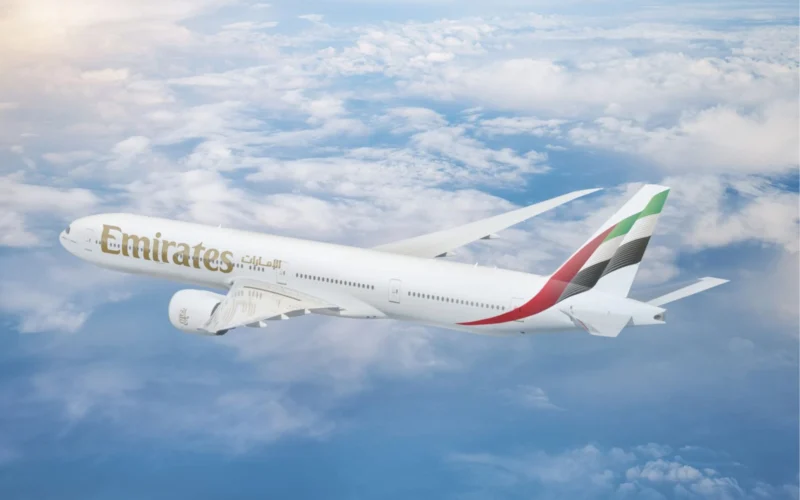 Emirates passenger jet flying above fluffy clouds under a blue sky with its wing visible