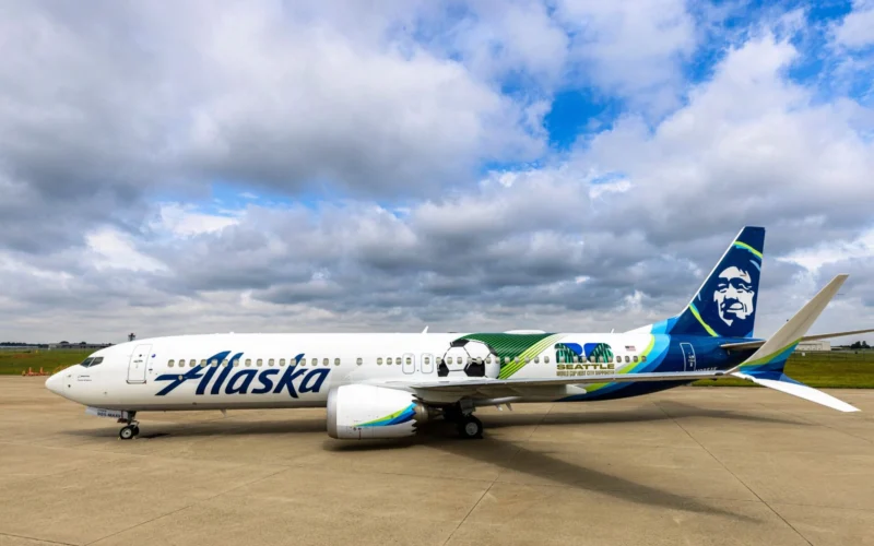 White Alaska Airlines jet on the tarmac with a colorful Seattle World Cup City liverie and tail art under a cloudy sky