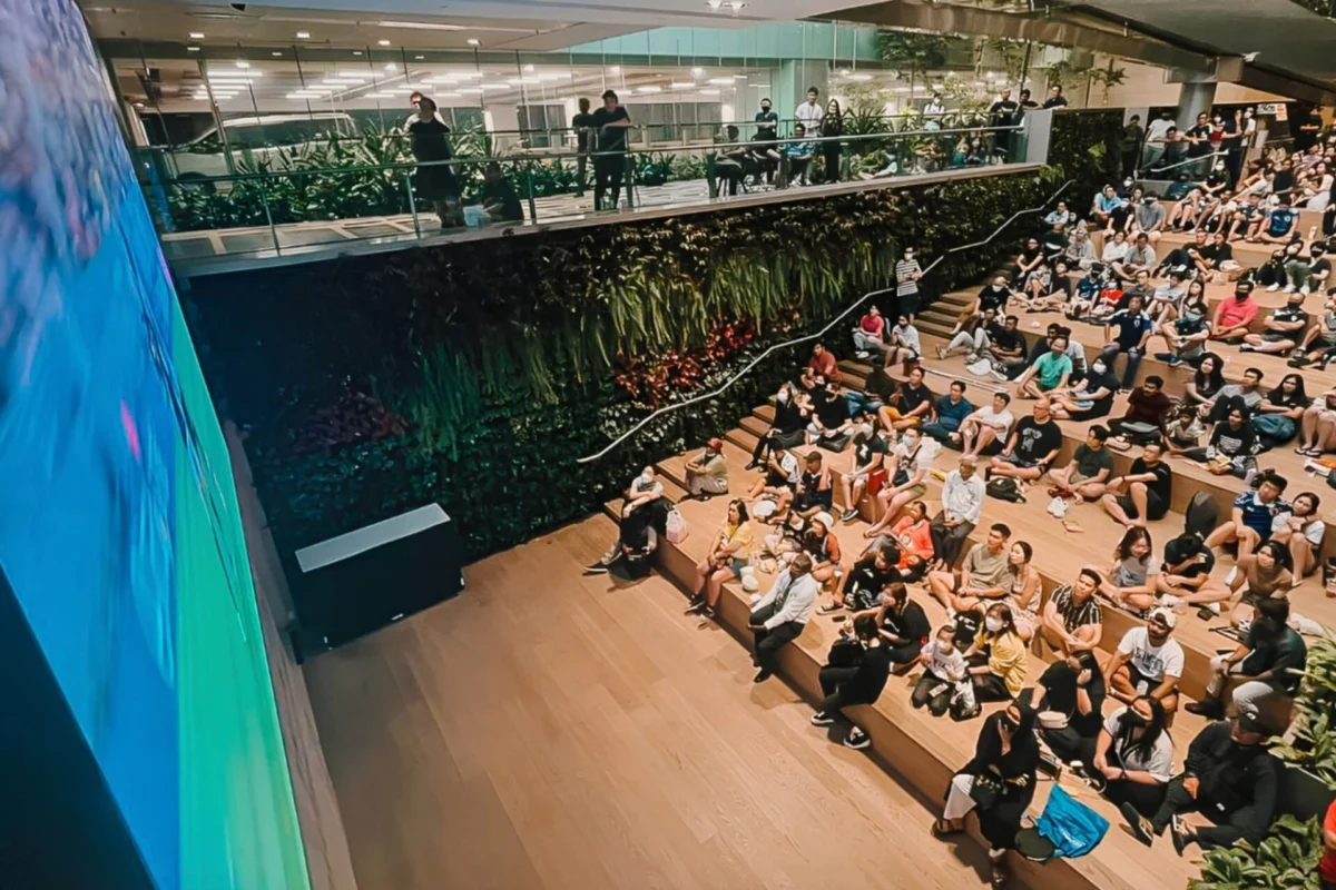 Large audience seated on tiered wooden steps in a modern indoor venue facing a blue curved stage wall in the foreground