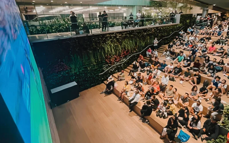 Large audience seated on tiered wooden steps in a modern indoor venue facing a blue curved stage wall in the foreground