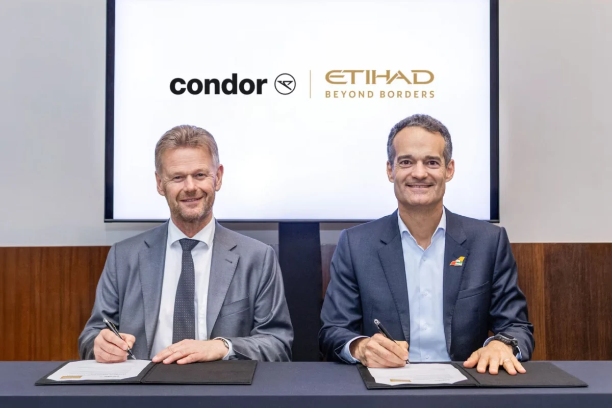 Two men in suits sign documents at a desk with Condor and Etihad Beyond Borders logos on a screen behind them