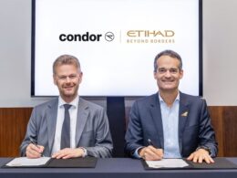 Two men in suits sign documents at a desk with Condor and Etihad Beyond Borders logos on a screen behind them
