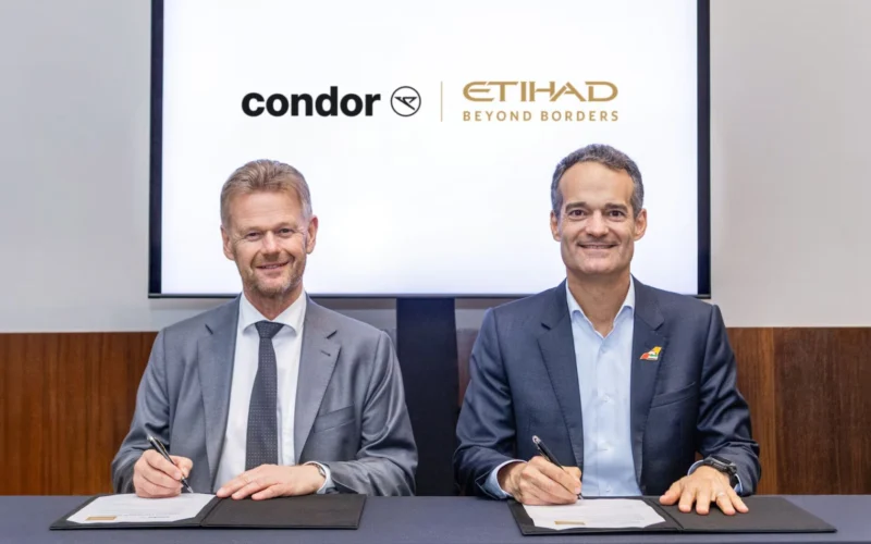 Two men in suits sign documents at a desk with Condor and Etihad Beyond Borders logos on a screen behind them