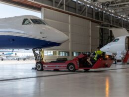 Large white and blue jet on a red maintenance vehicle inside a hangar with a technician in a hi vis vest nearby another jet with stairs in the background