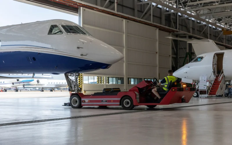 Large white and blue jet on a red maintenance vehicle inside a hangar with a technician in a hi vis vest nearby another jet with stairs in the background