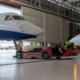 Large white and blue jet on a red maintenance vehicle inside a hangar with a technician in a hi vis vest nearby another jet with stairs in the background
