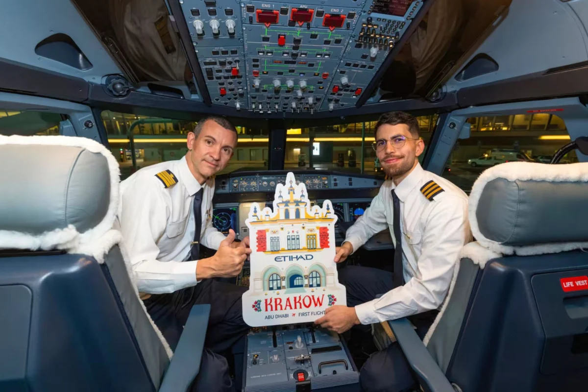 Etihad now flies to Krakow and Palma de Mallorca Two pilots in uniform sit in an airplane cockpit smiling and holding a Krakow promotional cutout showing ABU DHABI FIRST FLIGHT