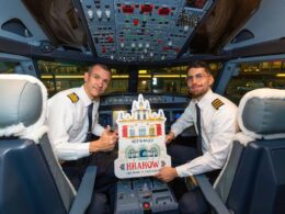 Two pilots in uniform sit in an airplane cockpit smiling and holding a Krakow promotional cutout showing ABU DHABI  FIRST FLIGHT