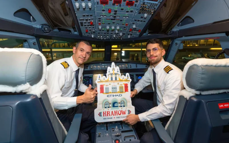 Two pilots in uniform sit in an airplane cockpit smiling and holding a Krakow promotional cutout showing ABU DHABI  FIRST FLIGHT