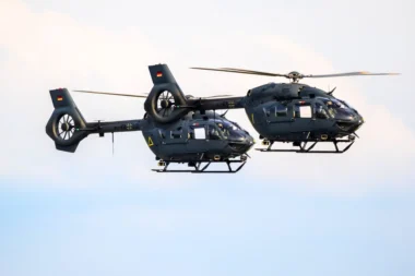 Two dark gray military helicopters flying side by side against a blue sky