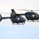 Two dark gray military helicopters flying side by side against a blue sky