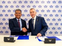 Two men in suits shake hands at a signing ceremony against an IATA backdrop with documents on the table behind them