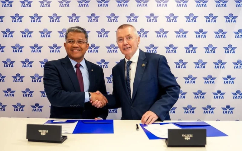 Two men in suits shake hands at a signing ceremony against an IATA backdrop with documents on the table behind them
