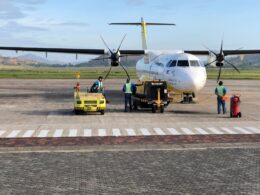 White twin engine ATR style aircraft parked on the tarmac while ground crew in high visibility vests operate yellow support equipment nearby