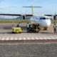 White twin engine ATR style aircraft parked on the tarmac while ground crew in high visibility vests operate yellow support equipment nearby