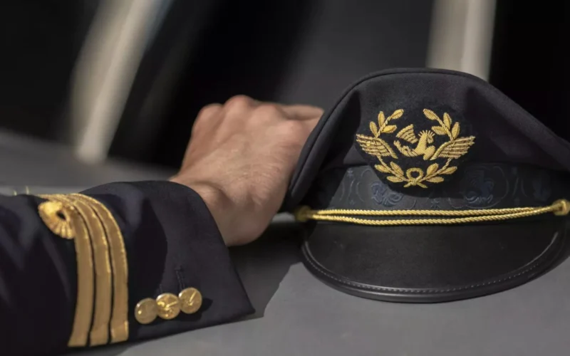 Pilots hand rests on a dark captains hat with gold embroidery and braid detail on a table