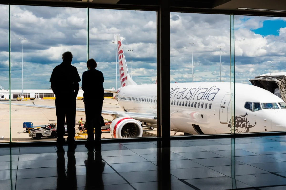 Virgin Australia resumes Doha flights from Melbourne Airport Two people silhouetted at a gate watching a large white commercial airplane on the tarmac outside under a cloudy sky