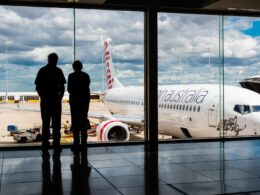 Two people silhouetted at a gate watching a large white commercial airplane on the tarmac outside under a cloudy sky