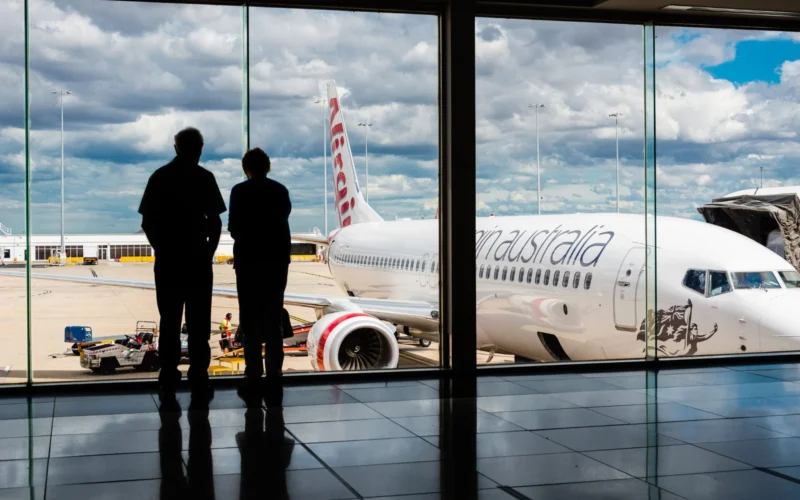 Two people silhouetted at a gate watching a large white commercial airplane on the tarmac outside under a cloudy sky