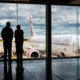 Two people silhouetted at a gate watching a large white commercial airplane on the tarmac outside under a cloudy sky