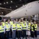 Group of airline staff in yellow safety vests posing in a hangar with a large white plane behind them
