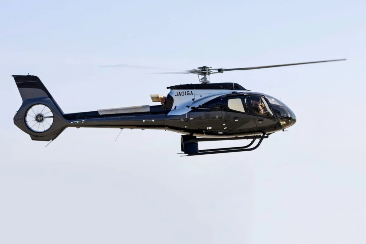 Side view of a sleek black helicopter in flight with spinning rotor blades against a pale blue sky JAOIGA markings on the fuselage landing gear visible underbody