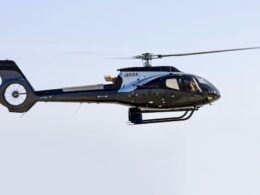 Side view of a sleek black helicopter in flight with spinning rotor blades against a pale blue sky JAOIGA markings on the fuselage landing gear visible underbody