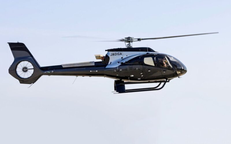 Airbus delivers ACH130 Aston Martin Edition to NOT A HOTEL Side view of a sleek black helicopter in flight with spinning rotor blades against a pale blue sky JAOIGA markings on the fuselage landing gear visible underbody
