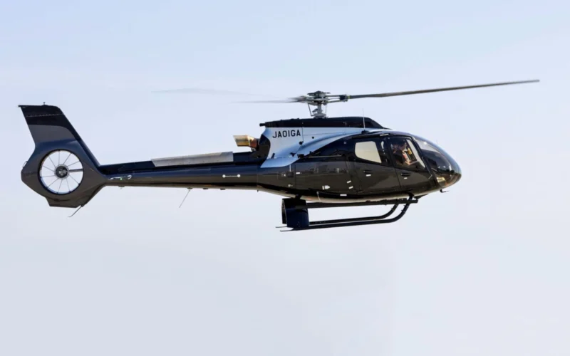 Side view of a sleek black helicopter in flight with spinning rotor blades against a pale blue sky JAOIGA markings on the fuselage landing gear visible underbody