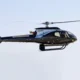 Side view of a sleek black helicopter in flight with spinning rotor blades against a pale blue sky JAOIGA markings on the fuselage landing gear visible underbody
