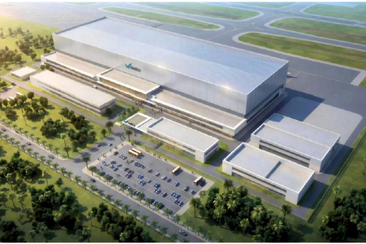 Aerial view of a large modern industrial complex with a long silver hangar smaller adjacent buildings and a parking lot lined with palm trees beside an airfield