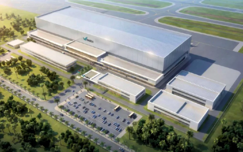 Aerial view of a large modern industrial complex with a long silver hangar smaller adjacent buildings and a parking lot lined with palm trees beside an airfield