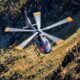 A helicopter flying over a rocky mountain landscape viewed from above with spinning rotors and a grassy valley below during daylight