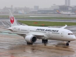 Front half of a Japan Airlines plane on a wet tarmac with a red JAL tail logo and airport buildings in the rainy background