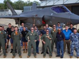 Group of pilots and crew in green flight suits and blue uniforms posing in front of a gray fighter jet on a tarmac