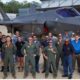 Group of pilots and crew in green flight suits and blue uniforms posing in front of a gray fighter jet on a tarmac