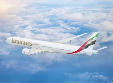 Emirates passenger jet in flight above the clouds, tail fin with red, green, white, and black stripes.