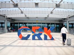 Colorful large letters spelling GRK on a sunny plaza outside a modern glass front building a man with a rolling suitcase walks by