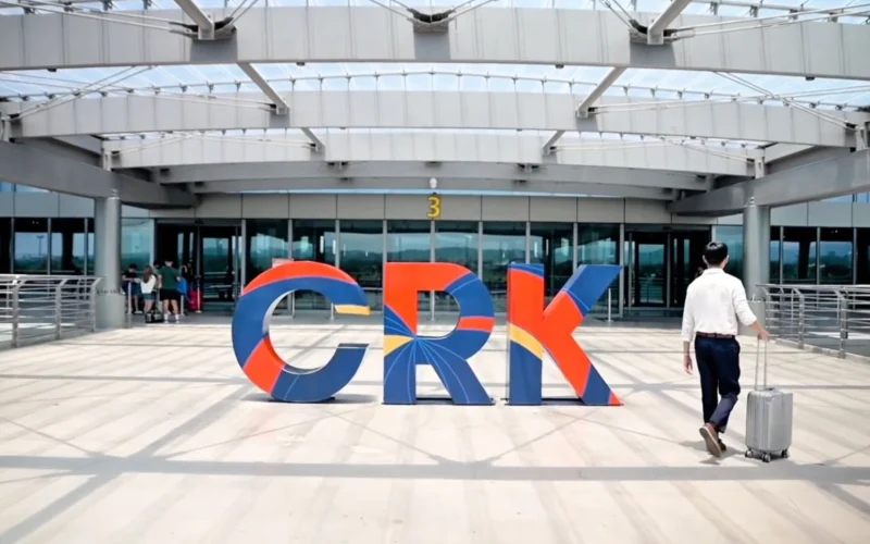 Colorful large letters spelling GRK on a sunny plaza outside a modern glass front building a man with a rolling suitcase walks by